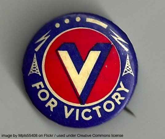 an old pin with a white V on a red background, while around the edge is written "FOR VICTORY"
