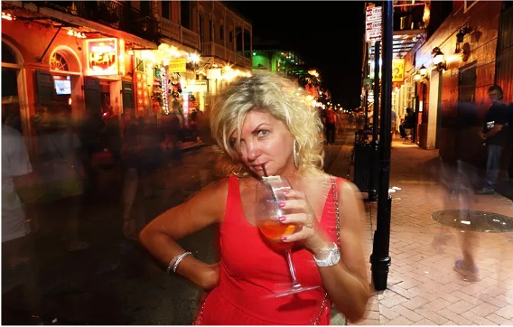 a very drunk woman in a red dress stands on bourbon st. in new orleans sipping a drink and looking belligerent