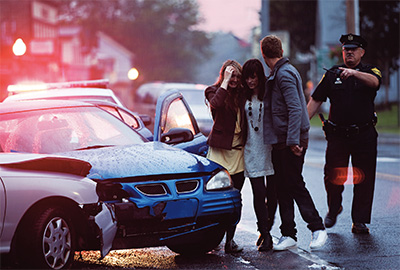 At dawn, three adolescents cry near a car wreck as a police officer looks on